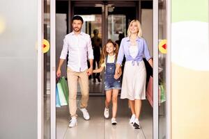 A family, consisting of a father, a mother, and a young daughter, walks out of a shopping mall, carrying shopping bags. The parents are holding hands with their daughter, and they are all smiling photo