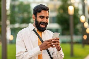 Indian bearded man is happily engaged with his smartphone while standing in a lush green park during a sunny afternoon. Lamps and modern buildings are visible in the blurry background photo
