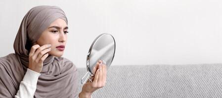Problem Skin Concept. Muslim Girl In Hijab Looking In Round Mirror And Touching Face, Concerned About Wrinkle Or Pimple, Closeup photo