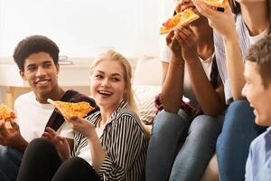A cheerful group of young friends sits comfortably on the floor and sofa in a well-lit living room, sharing slices of pizza and engaging in lively conversation photo