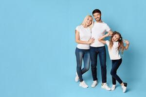 A family of three stands in front of a light blue background. The mother and father are standing close together, while the daughter stands slightly to the right, copy space photo
