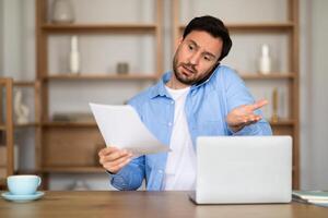 A perplexed man is seated at a wooden desk, talking on phone, holding a document and gesturing questioningly as he gazes toward the camera, with an open laptop in front of him indicating work photo