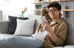 Enjoying Weekend. Handsome Asian Man In Glasses Using Smartphone While Sitting Leaning On Couch At Home, Happy Guy Relaxing In Living Room, Browsing Internet Or Messaging With Friends, Selective Focus photo