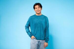Asian guy stands with his hands in his pockets in front of a bright blue wall. He gazes ahead with a neutral expression under the clear sky. photo