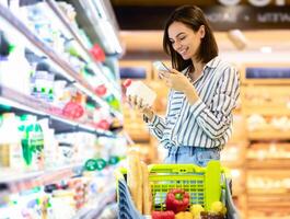 Smiling young woman taking dairy products from shelf in the supermarket, holding bottle and smartphone, scanning bar code on product through mobile phone, walking with trolley cart photo