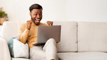 Yes. Excited Afro Man Using Laptop Computer Shaking Fists Celebrating Success Sitting On Sofa Indoor. Empty Space photo