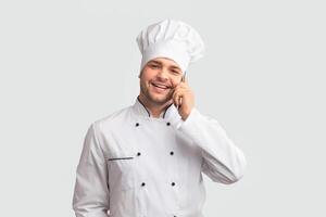 Culinary Services. Smiling Chef Cook Man In Uniform Talking On Phone Holding Clock Standing On White Studio Background. Panorama photo