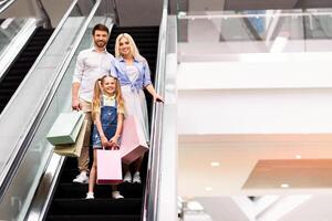 A happy family, a father, mother, and young daughter, are riding up an escalator in a modern shopping mall. They are all smiling and holding shopping bags, copy space photo