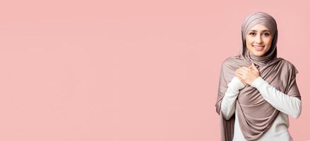 Thankful islamic girl in hijab with hands on chest expressing gratitude and looking at camera over pink background with empty space photo