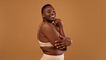 Self Acceptance, Love And Care. Portrair of half-turned smiling curvy African American woman with perfect skin and closed eyes embracing herself isolated on brown studio background, free copy space photo