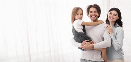Happy Father And Mother Posing With Their Cute Little Daughter On Hands, Standing Near Window At Home, Empty Space photo