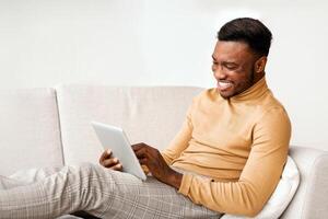 Happy African American Guy Using Tablet Computer Browsing Internet Sitting On Couch At Home. Selective Focus photo