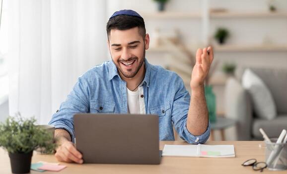 distante comunicación concepto. retrato de contento judío masculino en kipá y casual camisa haciendo llamada sentado a escritorio a hogar oficina y hablando con colegas, estudiantes o familia, explicando foto