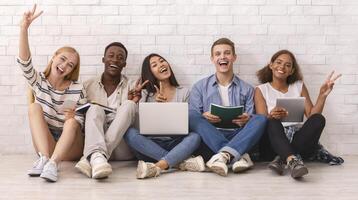 Group of happy multiracial students studying for university exams, cheerfully gesturing at camera, white brick wall background photo