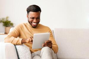 Smiling Black Guy Using Tablet Browsing Internet Enjoying Weekend Sitting On Couch Indoor. Selective Focus photo
