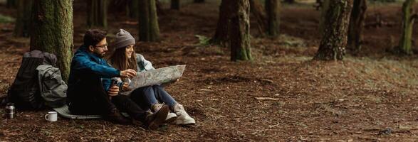 A young couple sits on the forest floor, surrounded by towering trees and fallen leaves. They are studying a map, likely planning their next adventure, copy space photo