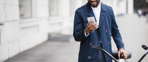 An African American businessman is commuting to his office by bicycle, using his phone. The mood is dynamic and contemporary, emphasizing multitasking and active transportation, cropped photo