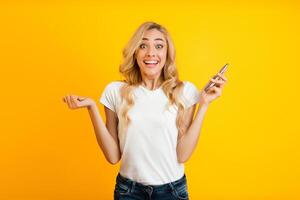 A blonde woman stands in front of a bright yellow background with a surprised expression on her face, holding a smartphone in one hand, and her other hand outstretched in a gesture of disbelief photo