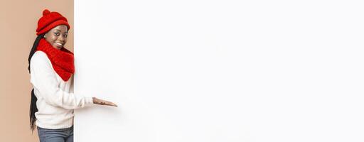 Empty Space For Advertising. Black cheerful woman in red knitted set directing with palm to empty white board, beige background, panorama photo