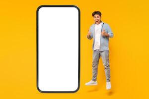 A young man jumps in the air in front of a large smartphone with a blank white screen, giving a thumbs up with both hands and a big smile on his face. The background is a bright yellow photo
