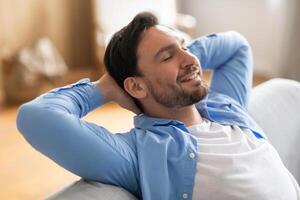 A man is seated on a couch, leaning back with his hands on his head. He appears relaxed and contemplative as he takes a moment to unwind at home. photo