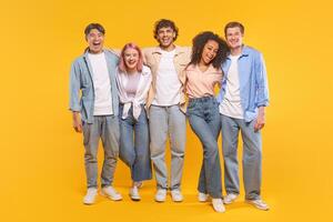 Five young adults are standing together in front of a yellow background. They are all smiling and laughing, and they are dressed in casual clothing. The group seems to be having a lot of fun photo