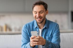 A man sits in a kitchen, wearing a blue button-up shirt and white undershirt, looking down at his smartphone with a wide grin. He is excited by what he is reading. photo