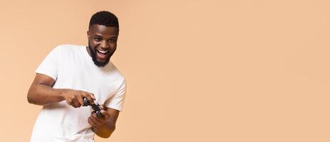 A young black man wearing a white t-shirt is playing a game with a controller in his hands. He is smiling and looking happy as he plays. The background is a light peach color. photo