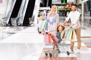 A young girl sits in a shopping cart being pushed by her father, while her mother walks behind with shopping bags. The family is smiling and laughing as they navigate the modern mall photo