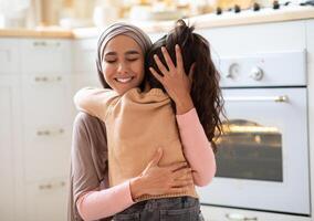 Happy Mother's Day. Little Girl Cuddling Tight Her Muslim Mom In Kitchen At Home, Expressing Love And Affection. Positive Islamic Woman In Hijab Hugging Her Child And Smiling, Closeup With Copy Space photo
