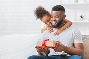 Black father sits on a couch with his daughter embracing him from behind. The father is opening a gift wrapped in brown paper and red ribbon. Both appear to be smiling and happy. photo