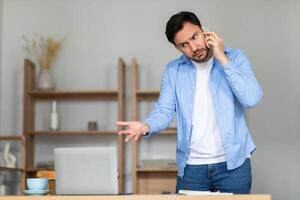 A man stands in his home office, with shelves in the background, gesturing with his hand while talking on a smartphone. He appears puzzled or concerned, possibly discussing an important matter photo