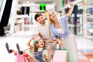 A family of three is shopping at a mall. The young girl is sitting in shopping cart while her parents take a selfie. The mother is holding the phone and smiling broadly. The father is standing behind photo