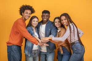 This image shows a diverse group of five friends stacking their hands together in a studio setting. They are all smiling and appear to be enjoying each others company. The background is bright yellow photo