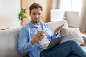A man sits on a couch with a laptop on his thighs, holding a smartphone in one hand while displaying a puzzled expression, appears to be encountering unexpected news or a perplexing situation photo