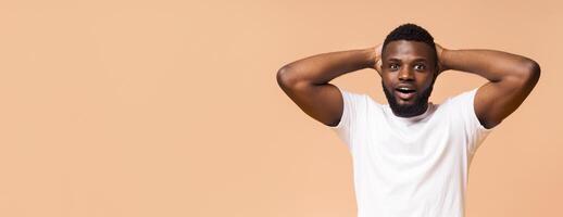 Black man with a beard and a white t-shirt standing in front of a tan background. He has his hands on his head and a shocked expression on his face. He looks like he is surprised or amazed, copy space photo