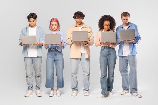 Five young adults are standing in a row in a studio setting. Each person is looking down at a laptop computer, their faces illuminated by the screen light photo