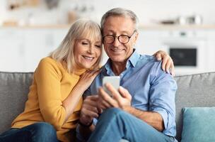 Happy Senior Couple Using Smartphone Browsing Internet Together Sitting On Couch At Home And Hugging. Cellphone Users Use Mobile Application. Older Spouses Texting On Phone. Communication Concept photo