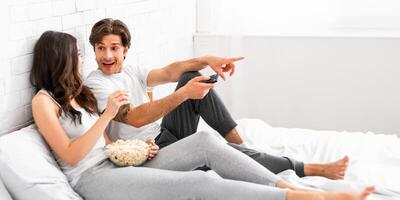 A young couple is comfortably sitting in bed together, watching television and enjoying a bowl of popcorn. The woman is smiling while the man is pointing at the TV and appears to be laughing. photo