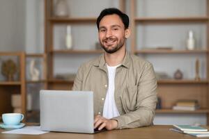 A man with a cheerful expression is seated at a desk with a laptop computer in front of him. He appears engaged in his work, typing on the keyboard and looking at the screen attentively. photo