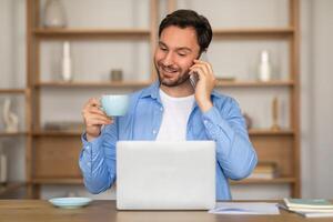 A man is engaged in a phone conversation while holding a cup of coffee in his other hand. He appears to be multitasking, balancing the phone against his ear while sipping from the steaming cup. photo