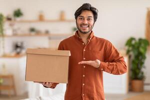 Arab man is holding a cardboard box in front of him with a big smile and his palm open toward the camera. The background is blurred and out of focus, suggesting a home or office environment. photo
