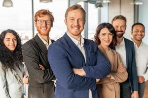 A group of diverse business professionals stands in an office setting, all smiling confidently and looking at the camera. They are wearing professional attire, man in the front has his arms crossed photo