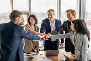 A group of five diverse business professionals stand around a conference table, smiling and stacking their hands on top of each other in a gesture of unity and teamwork. photo