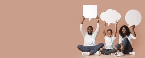 Diverse thoughts. Happy African american family of three holding empty speech bubbles over heads sitting on floor, yellow background with free space. photo