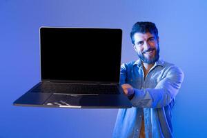 A cheerful man with a beard holds a laptop with a blank screen, showcasing it toward the camera. The setting features a blue gradient background and appears to be taken during the evening. photo