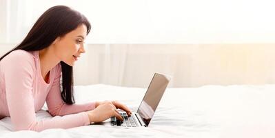 A woman reclining on a bed while actively engaged with a laptop computer, focusing on the screen and typing with hands on the keyboard, side view, panorama with copy space photo