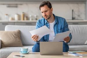 A man sits on a white couch in a living room, reviewing documents. He has a concerned look on his face as he carefully looks over the papers while sitting in front of an open laptop photo