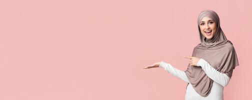 Beautiful muslim woman in hijab demonstrate something on her empty palm and pointing at it, standing over pink background, panorama with copy space photo