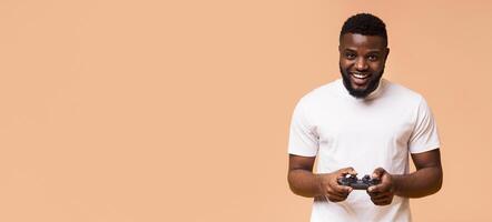 A smiling black man is shown holding a game controller in front of a peach-colored background. The man appears to be enjoying himself and is likely immersed in a game, copy space photo